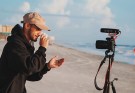 Mann som filmar seg sjølv med kamera på ein tripod på ei strand