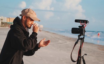 Mann som filmar seg sjølv med kamera på ein tripod på ei strand
